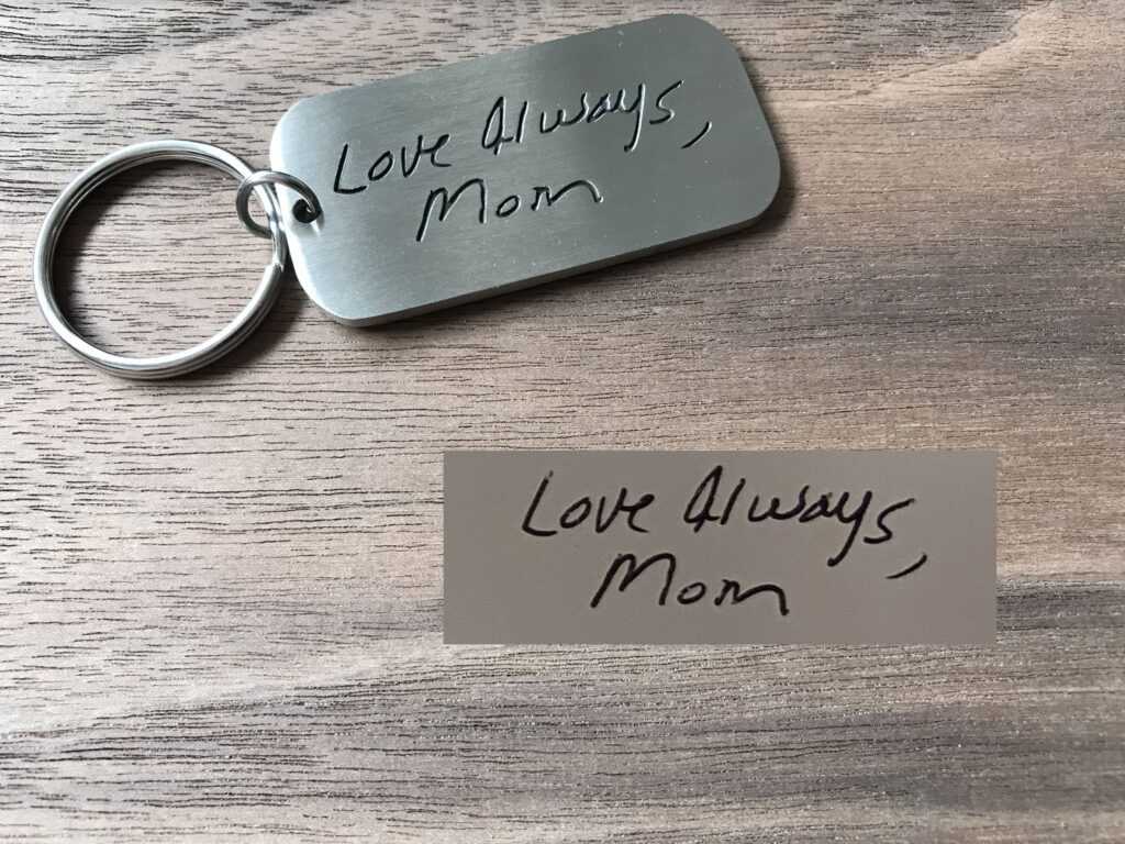 Silver keychain with engraved handwriting Love Always, Mom on a wooden surface.