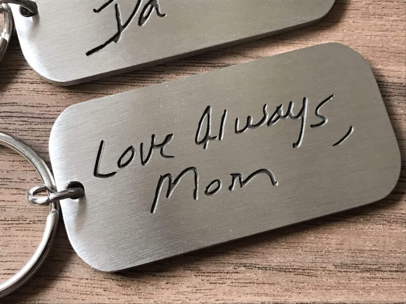 Engraved keychain with handwritten message Love Always, Mom on a metal tag, resting on a wooden surface.