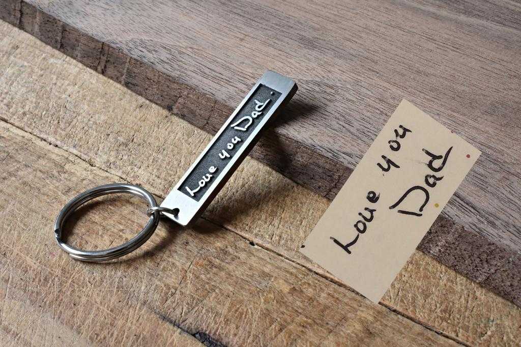 Keychain with Love you Dad engraving beside a handwritten note on a wooden surface. Perfect Father's Day gift.