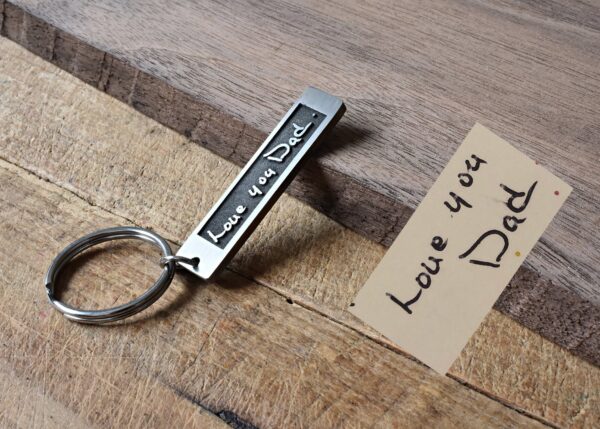 Keychain with Love you Dad engraving beside a handwritten note on a wooden surface. Perfect Father's Day gift.