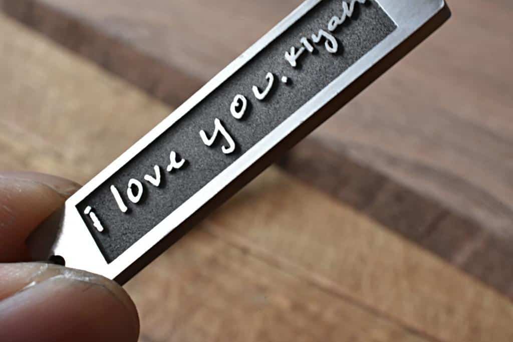 Engraved metal pendant reading I love you, with a finger holding it on a wooden background.