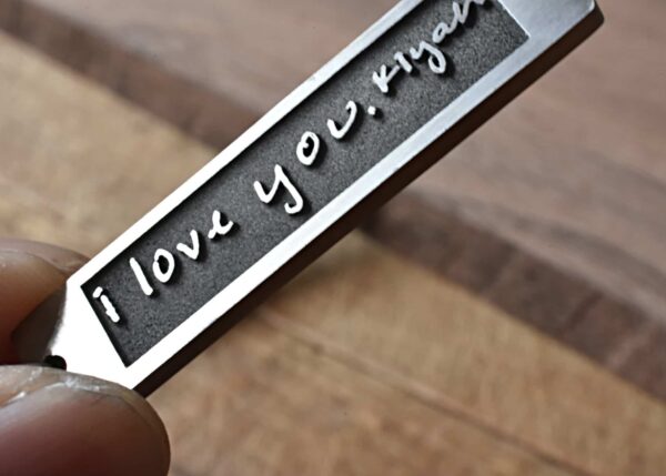 Engraved metal pendant reading I love you, with a finger holding it on a wooden background.