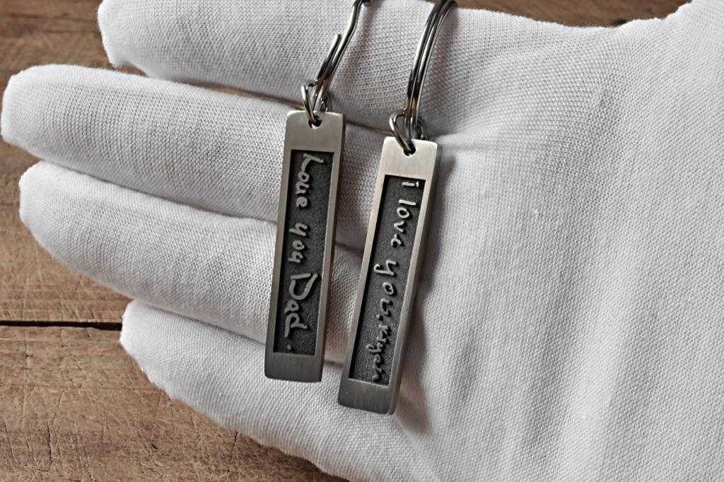 Keychains with Love you Dad and I love you, Mom engravings held in a white-gloved hand on wooden surface.