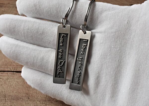 Keychains with Love you Dad and I love you, Mom engravings held in a white-gloved hand on wooden surface.