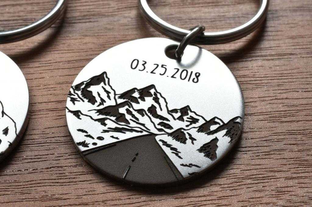 Round metal keychain with engraved mountains and date 03.25.2018, set on wooden surface.