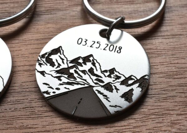 Round metal keychain with engraved mountains and date 03.25.2018, set on wooden surface.