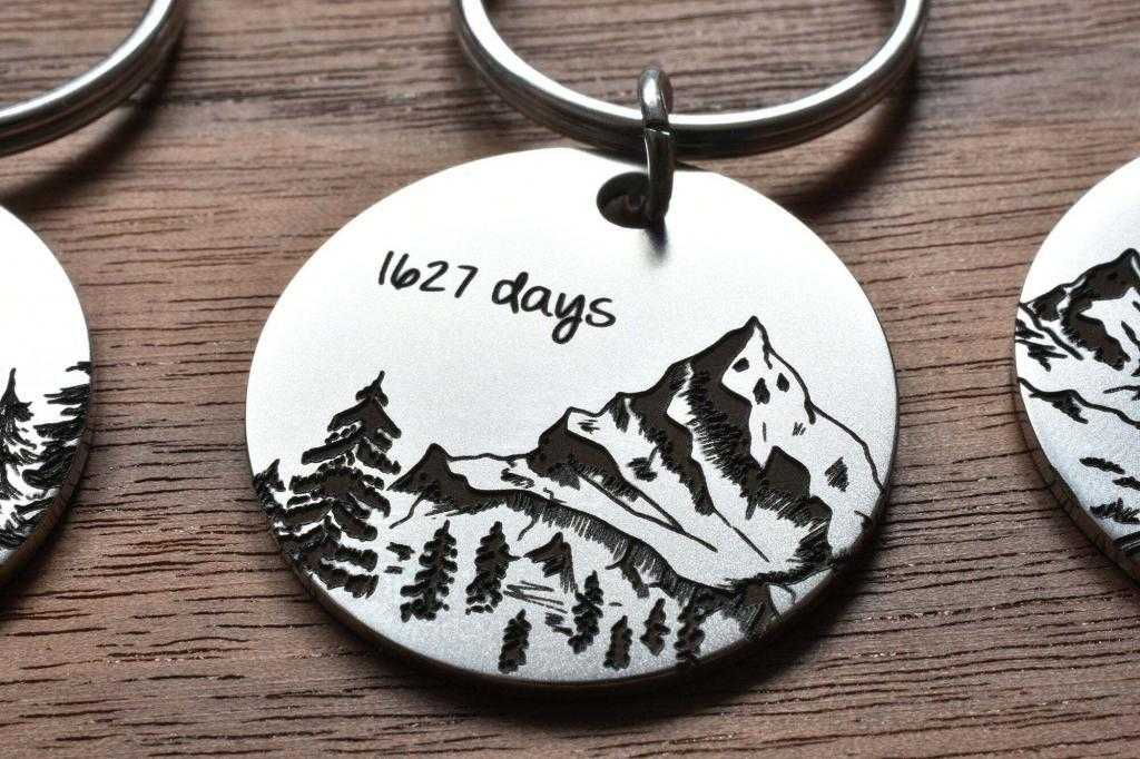 Engraved keychain with mountains and trees, marked with 1627 days, on a wooden surface.