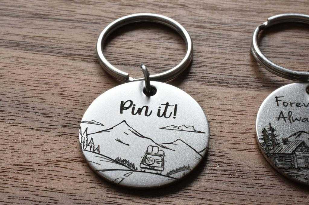 Keychain with Pin it! text, featuring a mountain scene and a car, lying on a wooden surface.