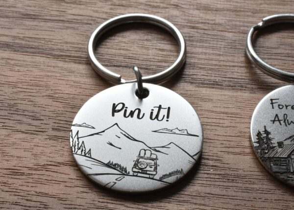 Keychain with Pin it! text, featuring a mountain scene and a car, lying on a wooden surface.
