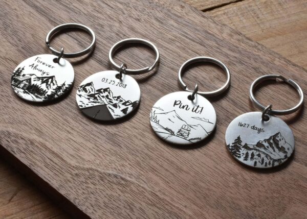 Four engraved round keychains on wooden surface, showcasing scenic mountain designs and custom text.