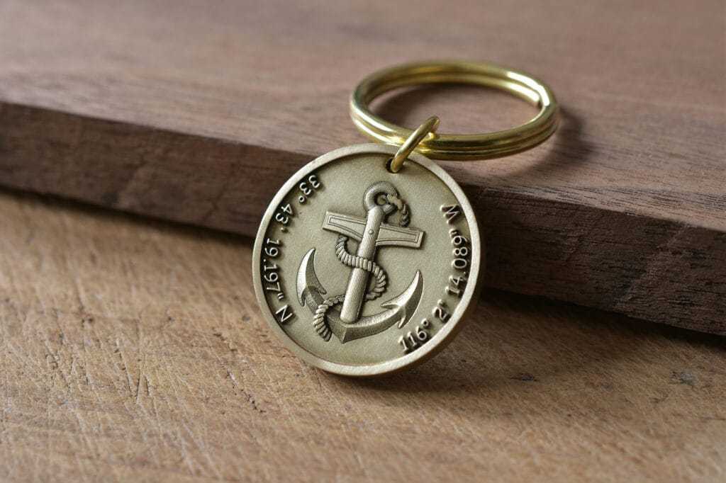 Gold keychain with anchor and engraved coordinates resting on wooden surface.