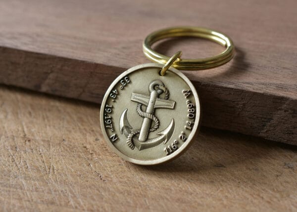 Gold keychain with anchor and engraved coordinates resting on wooden surface.