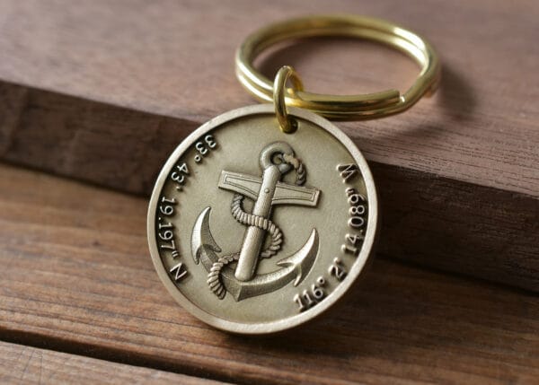 Gold keyring with engraved anchor and coordinates, resting on wooden surface, symbolizes nautical themes and travel.