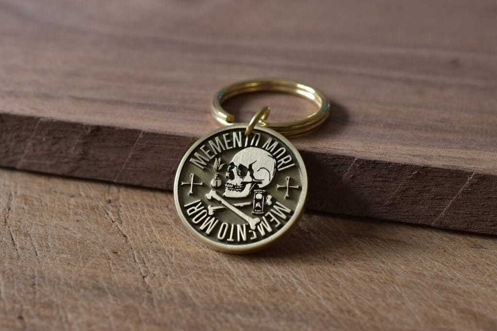 Gold keychain with Memento Mori design featuring a skull, crossbones, and hourglass on a wooden surface.