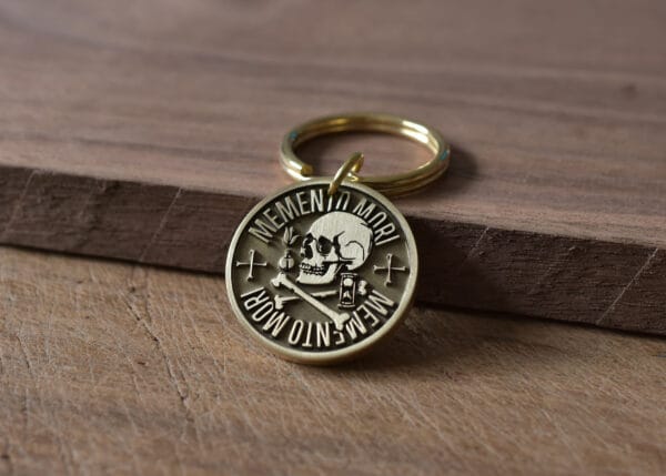 Gold keychain with Memento Mori design featuring a skull, crossbones, and hourglass on a wooden surface.