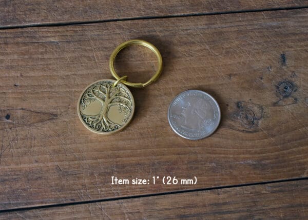 Gold tree of life keychain beside quarter for size comparison, on wooden table. Keychain size: 1 inch (26 mm).