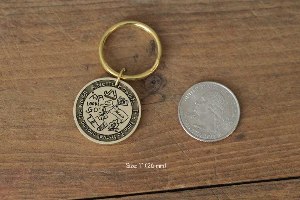 Round gold keychain with travel icons, text Adventure awaits, next to a US quarter for size comparison, on a wood surface.