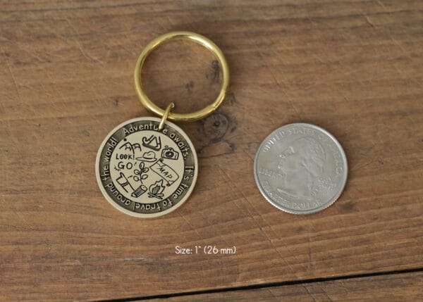Round gold keychain with travel icons, text Adventure awaits, next to a US quarter for size comparison, on a wood surface.