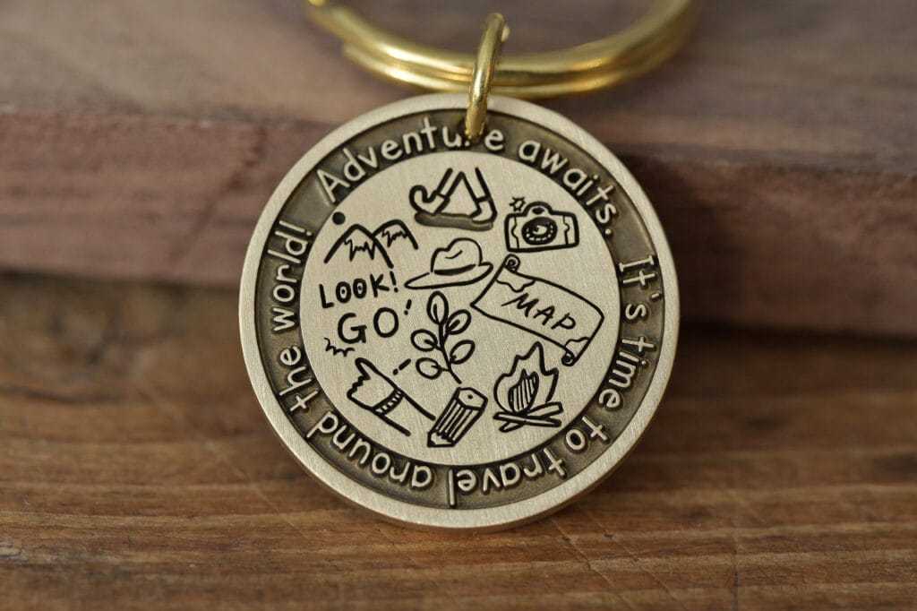 Gold keychain medallion with adventure-themed engravings: compass, camera, map, and inspiring text about travel.