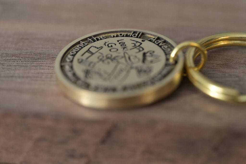 Close-up of a round, engraved brass keychain on a wooden surface, featuring travel-themed text and illustrations.