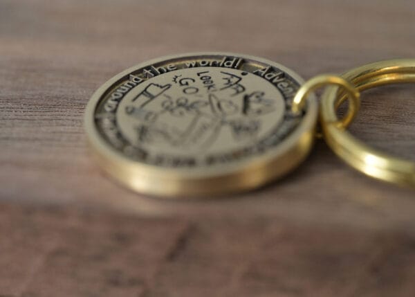 Close-up of a round, engraved brass keychain on a wooden surface, featuring travel-themed text and illustrations.