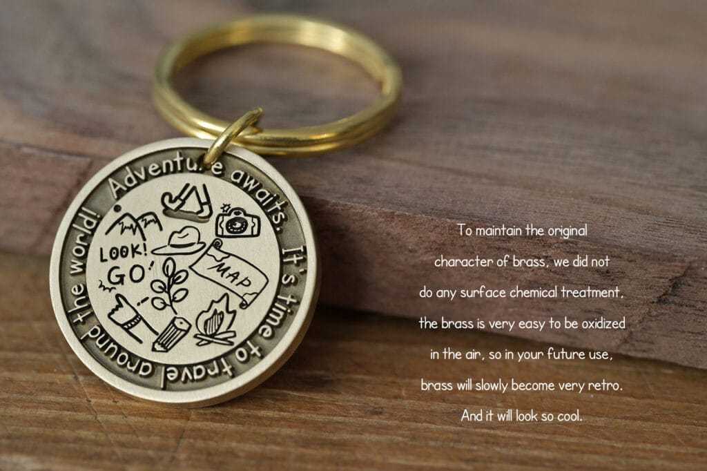 Brass keychain with adventure motifs, text: Adventure awaits. It’s time to travel around the world! on a wooden surface.