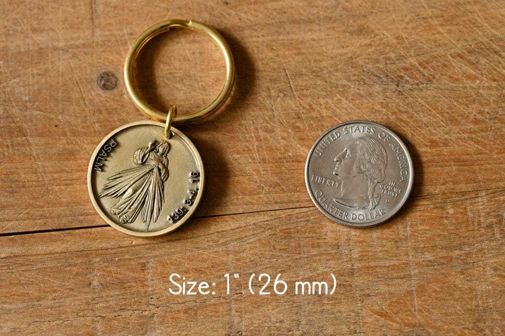 Gold keychain with a religious medallion next to a U.S. quarter for size comparison, on a wooden surface.