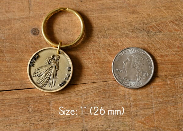 Gold keychain with a religious medallion next to a U.S. quarter for size comparison, on a wooden surface.