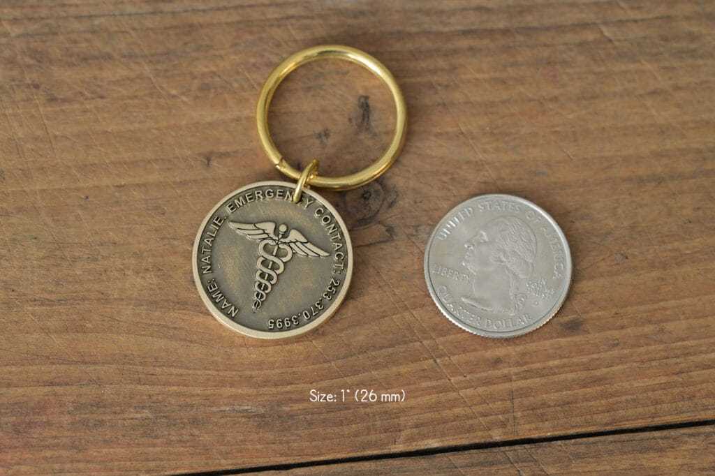 Gold medical alert keychain with Caduceus symbol next to a quarter on wooden surface, size comparison shown.