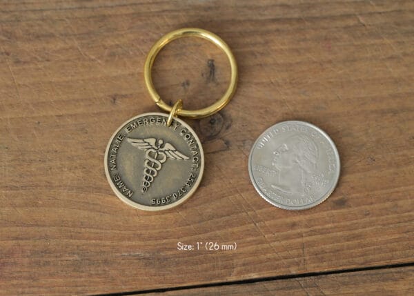 Gold medical alert keychain with Caduceus symbol next to a quarter on wooden surface, size comparison shown.