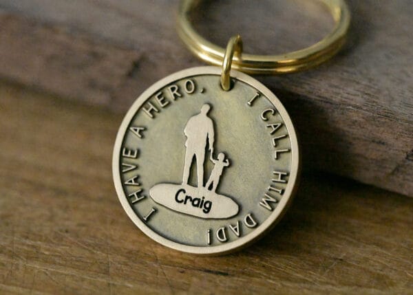 Gold keychain with engraved message I Have a Hero, I Call Him Dad, featuring silhouettes of a man and child.