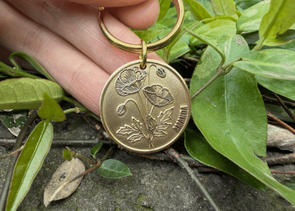 Gold keychain with poppy engraving and AMANDA text, held among green leaves on rocky ground.