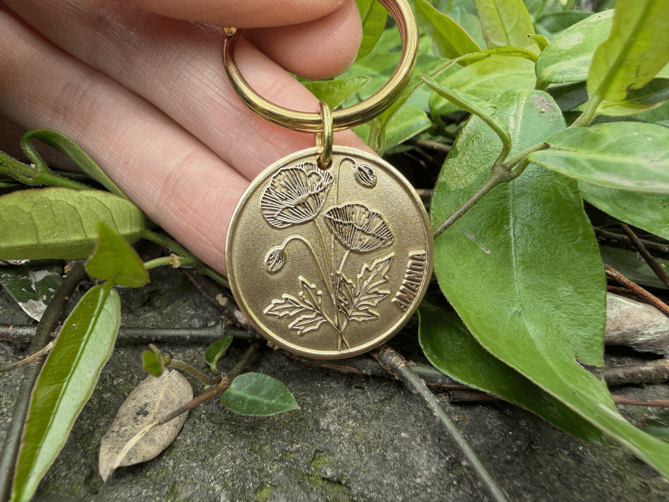 Gold keychain with poppy engraving and AMANDA text, held among green leaves on rocky ground.