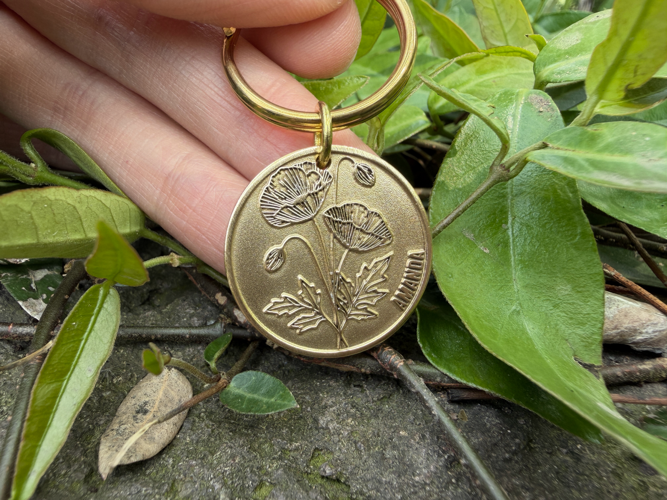 Gold keychain with poppy engraving and AMANDA text, held among green leaves on rocky ground.