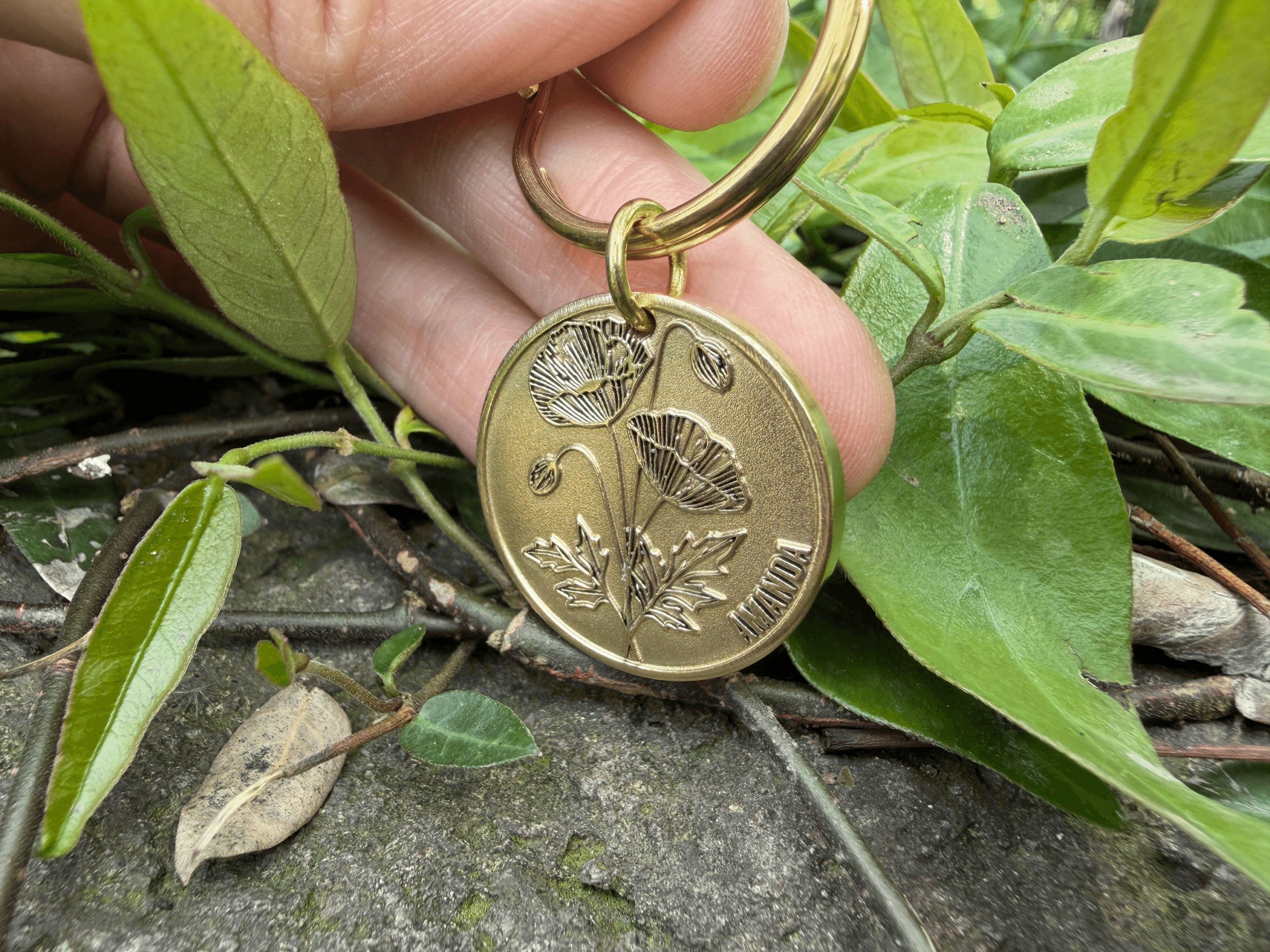 Gold coin keychain with engraved poppy flower design, held over leaves and pavement.