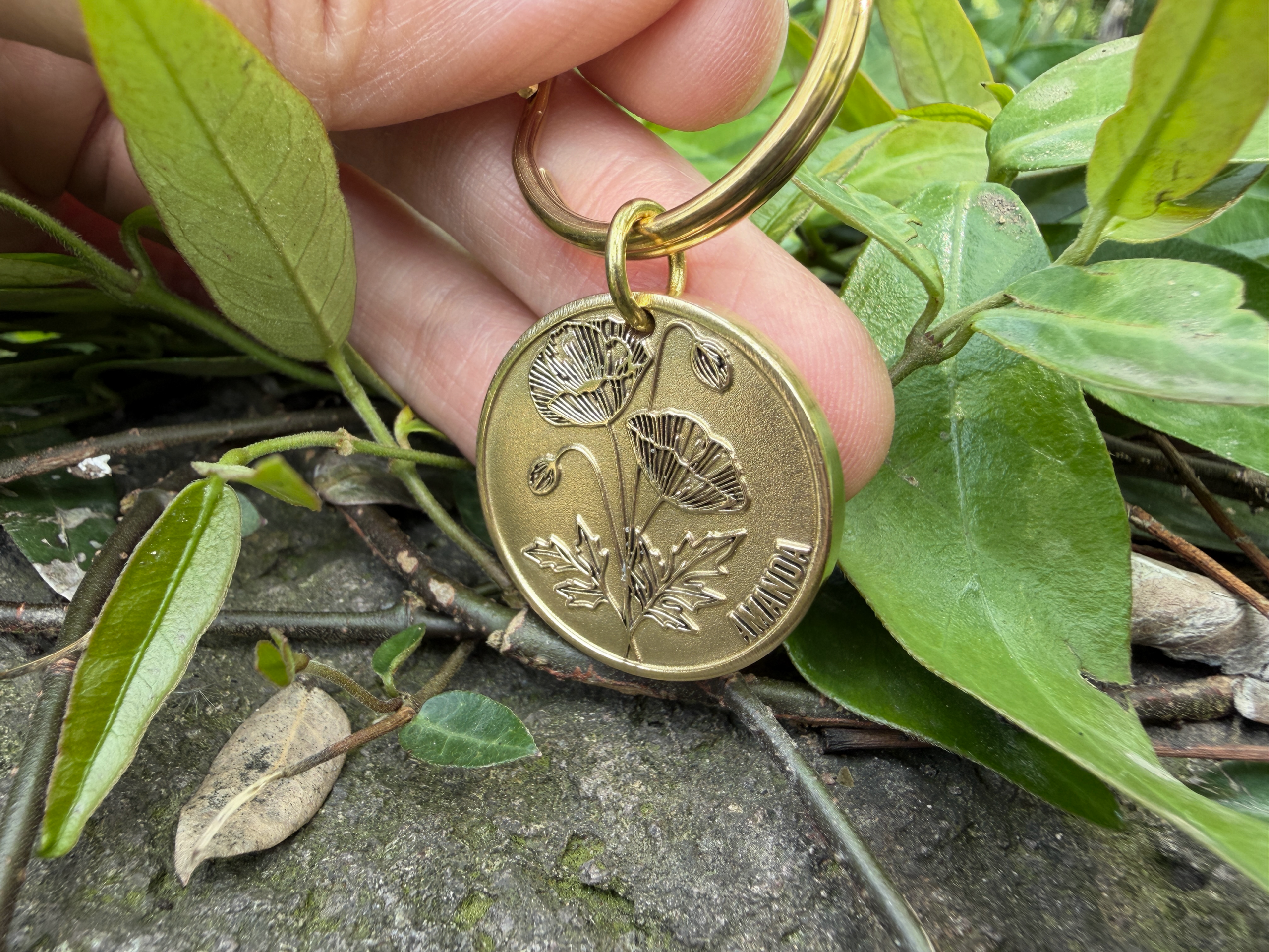 Gold coin keychain with engraved poppy flower design, held over leaves and pavement.