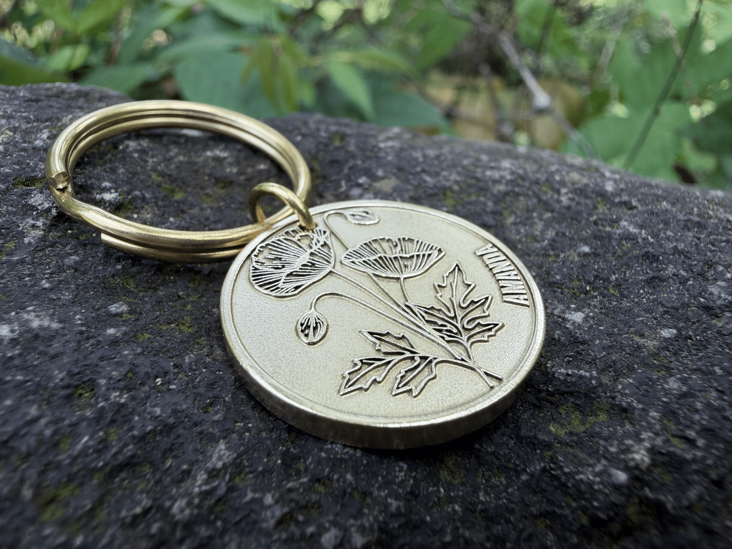 Keychain with engraved poppy flower and Amanda on a metal disc, placed on a rock surface.