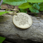 Golden floral keychain on driftwood surrounded by green leaves outdoors.