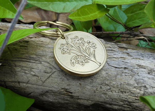 Golden floral keychain on driftwood surrounded by green leaves outdoors.