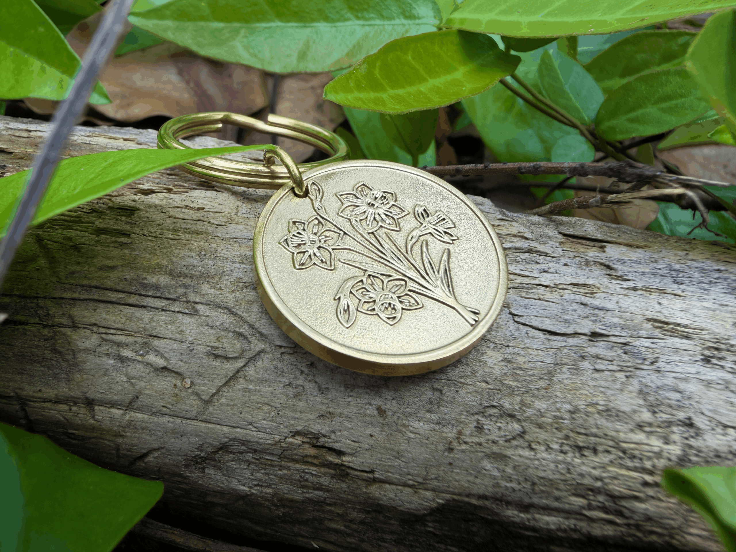 Golden floral keychain on driftwood surrounded by green leaves outdoors.