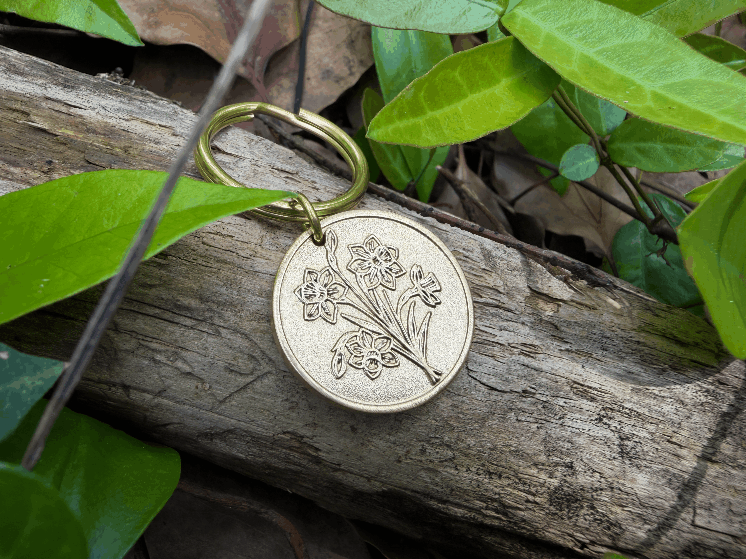 Floral coin keychain on a wooden log surrounded by green leaves in a natural setting.
