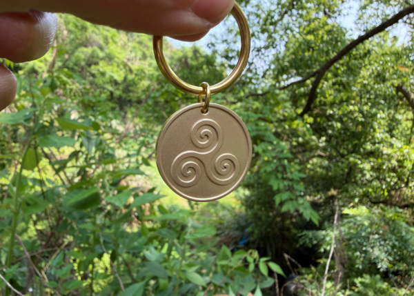 A person holds a keychain with a triskelion engraving, set against a lush green forest background.