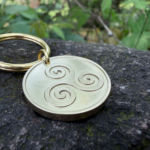 Gold keychain with a triskelion engraving on a stone surface; green foliage background.