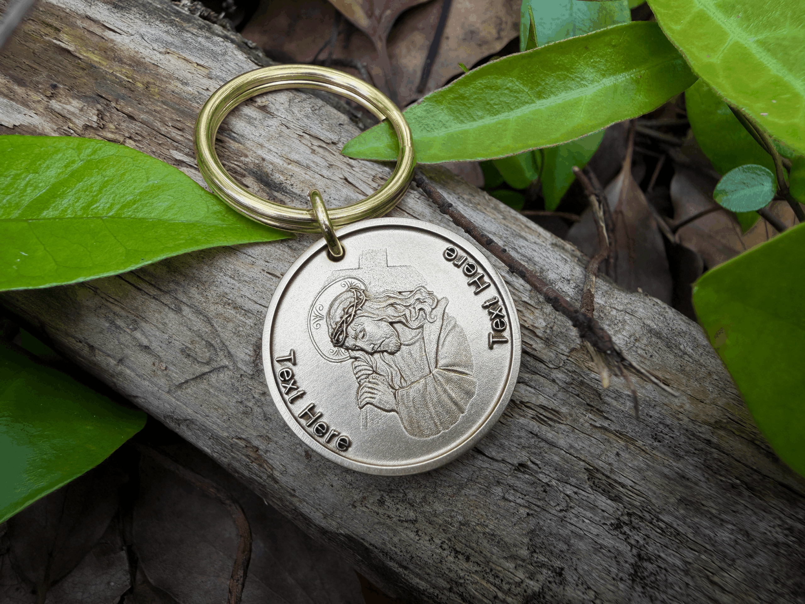 Keychain with engraving of Jesus praying, lying on wood with green leaves.