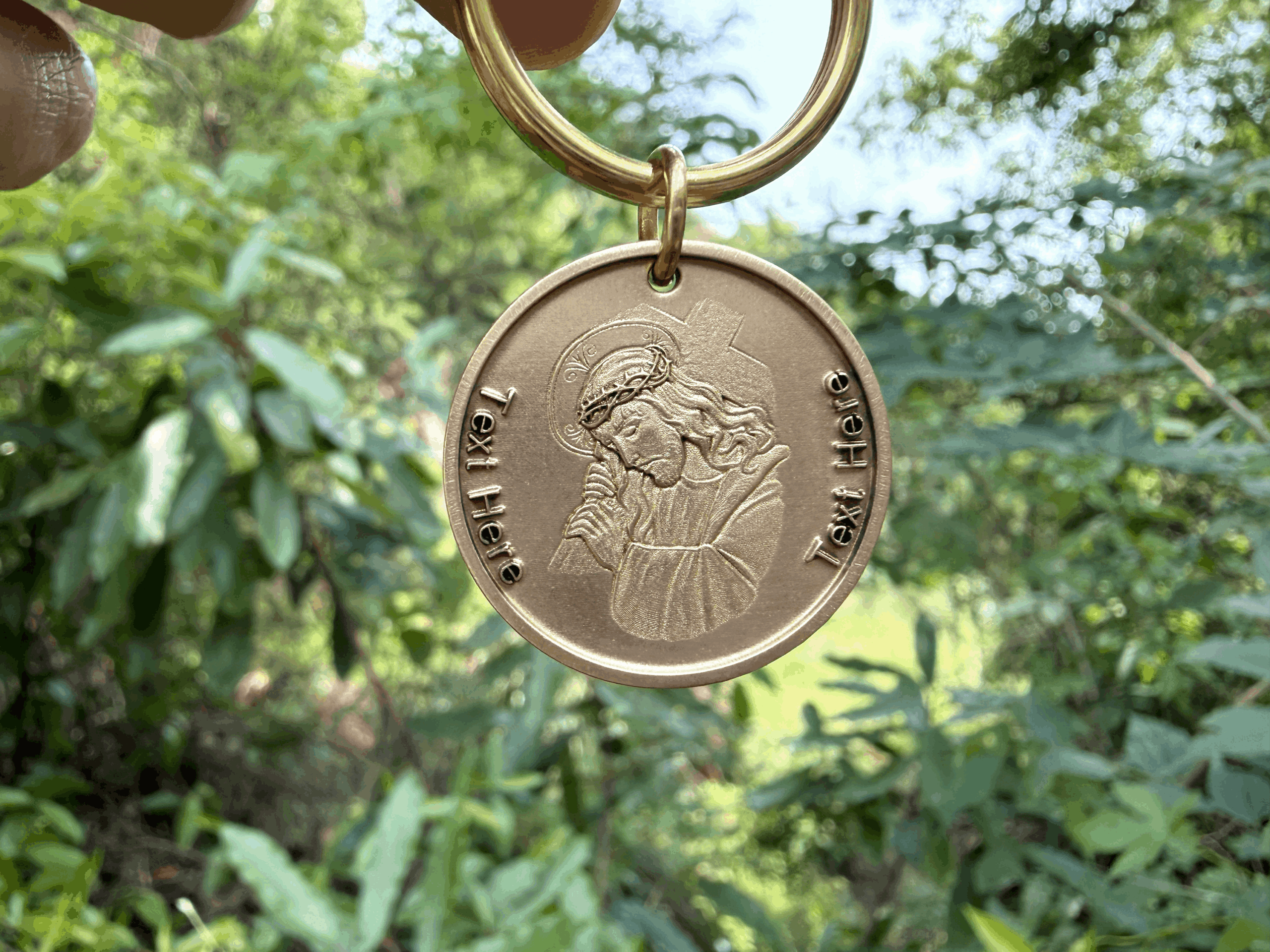 Gold medallion keychain with Jesus Christ engraving, held against a lush green background. Text unfinished.