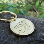 Golden keychain with Eye of Horus engraving on rock surface, surrounded by greenery in the background.