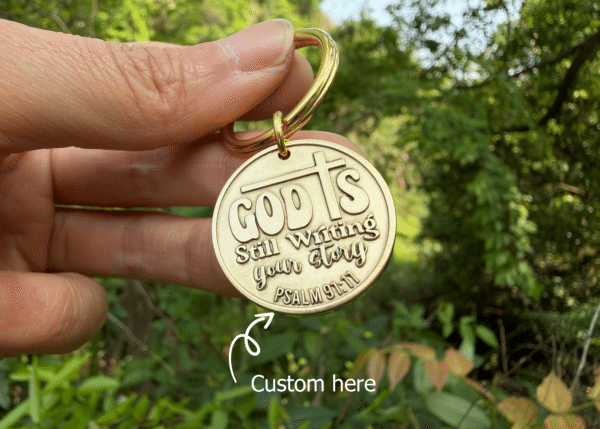 Keychain with God is still writing your story engraved, held in hand, nature background, Psalm 91:11 reference.