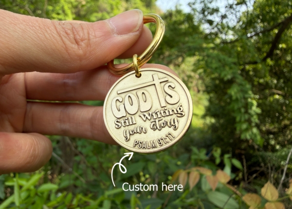 Keychain with God is still writing your story engraved, held in hand, nature background, Psalm 91:11 reference.
