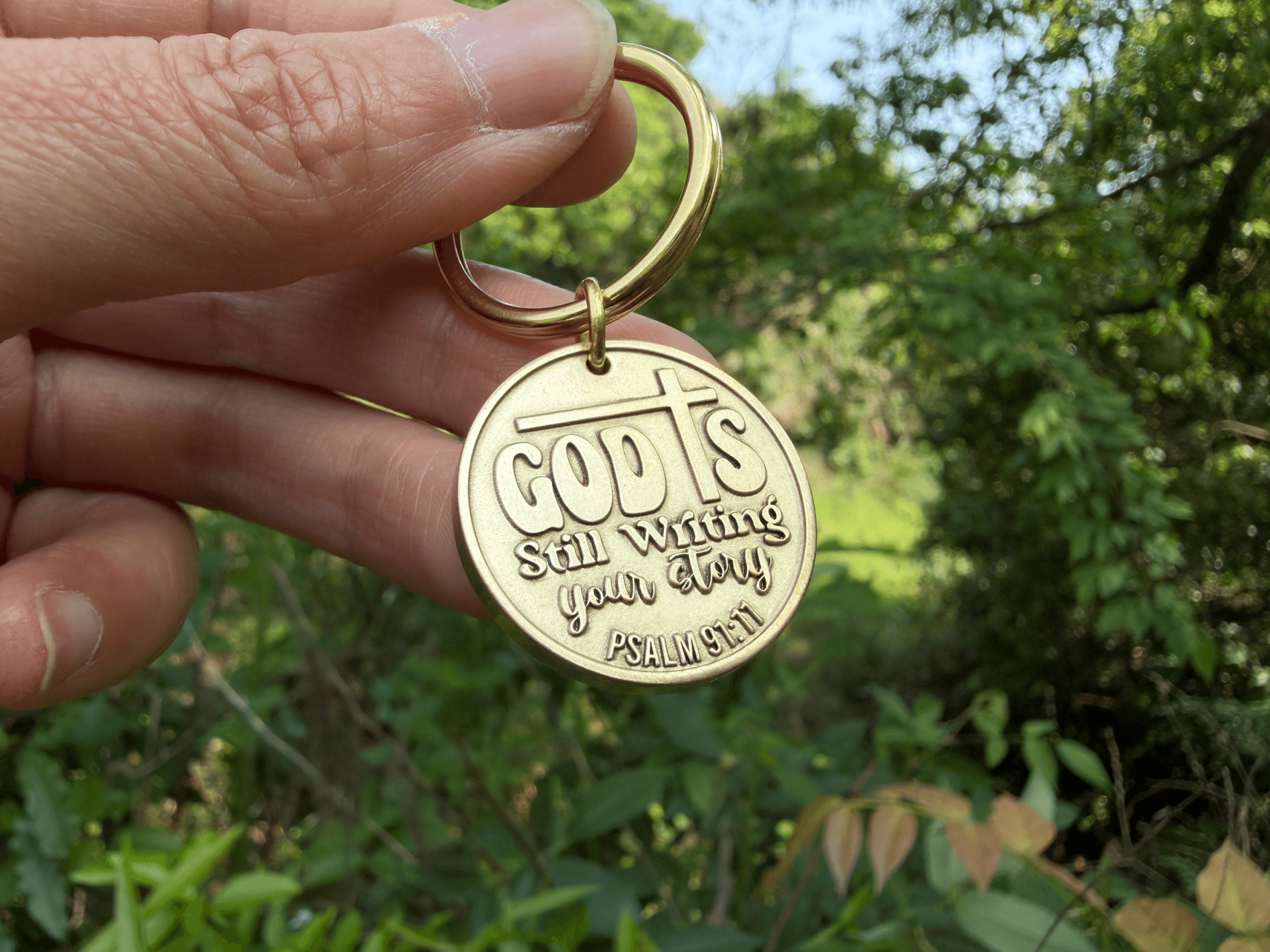 Gold keychain with engraving God's Still Writing Your Story, Psalm 91:11, held in nature background.