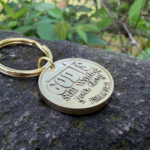 Gold keychain on rock engraved with God is still writing your story. Psalm 91:11 amidst greenery.